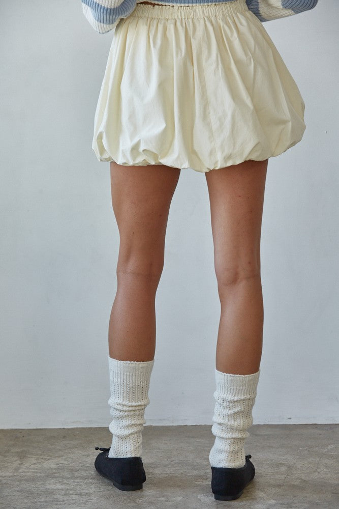 Person wearing a light-colored skirt, white socks, and black shoes against a plain background