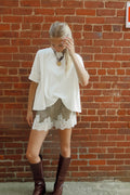 A woman stands against a red brick wall wearing a loose white short-sleeve top, light green shorts with white lace trim, and dark brown knee-high boots.