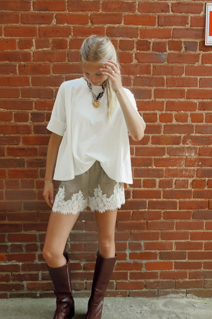 A woman stands against a red brick wall wearing a loose white short-sleeve top, light green shorts with white lace trim, and dark brown knee-high boots.