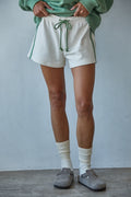 Person wearing white shorts, green sweater, and gray shoes against a plain background