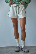 Person wearing white shorts, green sweater, and gray shoes against a plain background